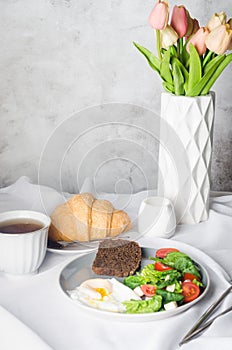 Salad in plate, egg, cup of coffee and croissant , fresh tulips in vase on clean white