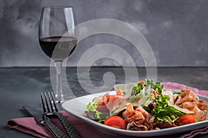 Salad with prosciutto and parmesan. Cutlery, knives, forks and pink napkin on the grey background. Glass of red wine