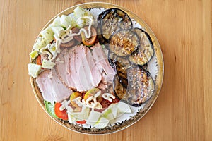 Salad on a round plate seen from above on a wooden table. The meal contains sliced ham, fried sliced aubergine, cucumber and