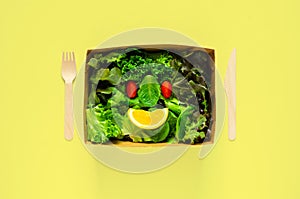 Salad set as smile face puts in disposable, compostable paper food box, fork and spoon on yellow background