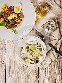 Salad with squid rings, mozzarella cheese, olives, capers and microgreens and baked vegetables with mushrooms in a white bowl.