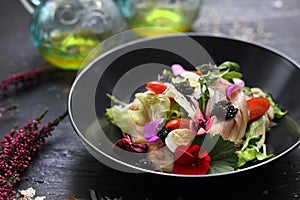 Salad with vegetables, fresh fish, caviar and quail egg, decorated with edible flowers. Salad in a black bowl, close up.