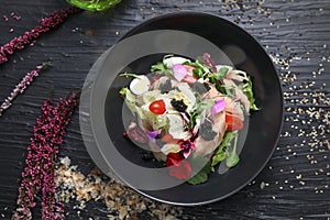 Salad with vegetables, fresh fish, caviar and quail egg, decorated with edible flowers. Salad in a black bowl, top view.