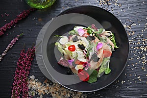 Salad with vegetables, fresh fish, caviar and quail egg, decorated with edible flowers. Salad in a black bowl, top view.