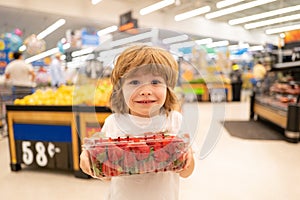 Sale, consumerism and people concept - happy little boy with food in shopping cart at grocery store with strawberry.