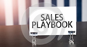 SALES PLAYBOOK sign on paper on dark desk in sunlight. Blue and white background