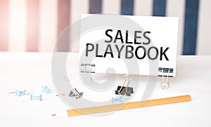 SALES PLAYBOOK sign on paper on white desk with office tools. Blue and white background
