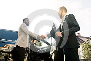 Sales situation in a car dealership, young Caucasian man dealer is shaking hands with a young African businessman, young