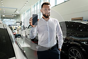 a salesman in a car dealership demonstrates the screen of a smartphone against the background of new cars