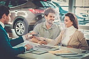 Salesman giving the key of the new car to a young couple at the dealership showroom.