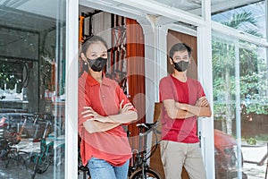 Salesman and saleswoman in mask stands leaning in front of the glass door with folded hands