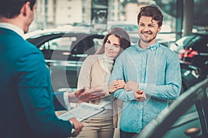 Salesman talking to a young couple at the dealership showroom.