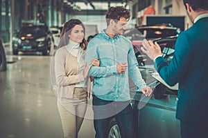 Salesman talking to a young couple at the dealership showroom.