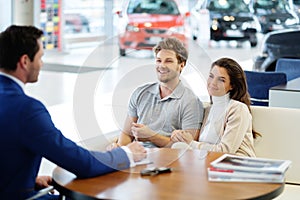 Salesman talking to a young couple at the dealership showroom.