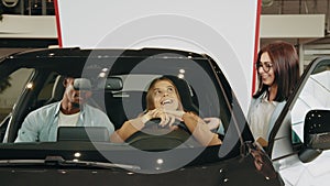 Saleswoman showing to diverse couple car at showroom. Beautiful multiracial couple sitting inside modern luxury car and