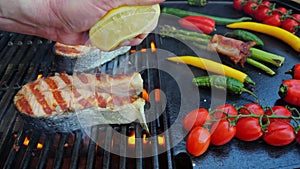 Salmon fish steak on grill with vegetables. Chef hand squeezes lemon on fish steak grilled. Slow motion. Close up
