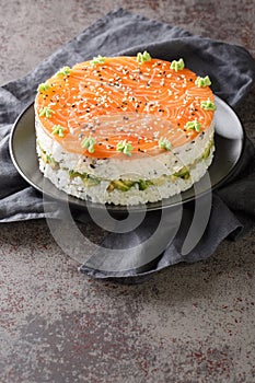 Salmon sushi cake with rice, avocado, cream cheese, seaweed, sesame seeds decorated with wasabi close-up in a plate. Vertical