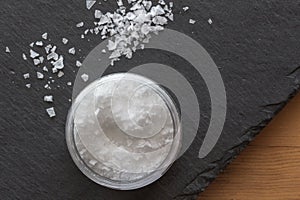 Salt in a container with salt flakes on black slate background on wooden table