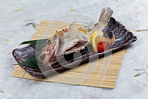 Salt-Grilled Snapper Shioyaki served with sliced lemon and tomato on stone plate on makisu Meal mat