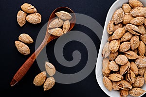 salted almonds in shell on white plate and spoon on black slate background top view