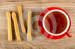 Salted bread sticks, cup with tea on saucer on wooden table. Top view