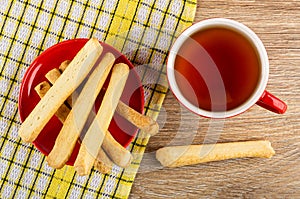 Salted bread sticks in saucer on napkin, cup with tea on wooden table. Top view