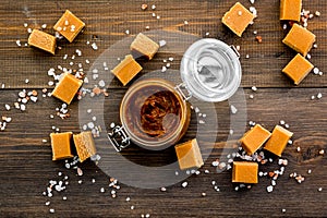 Salted caramel. Suace in glass jar and caramel cubes on dark wooden background top view