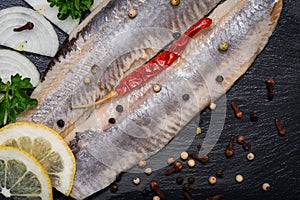 Salted Herring Fillets garnished with Yellow Onion, Lemon, Fresh Parsley and Dried Chili Pepper. Natural black stone background.