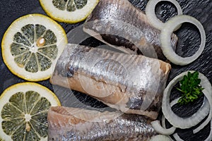 Salted Herring Fillets garnished with Yellow Onion, Lemon, Fresh Parsley and Dried Chili Pepper. Natural black stone background.