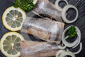 Salted Herring Fillets garnished with Yellow Onion, Lemon, Fresh Parsley and Dried Chili Pepper. Natural black stone background.