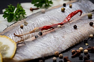 Salted Herring Fillets garnished with Yellow Onion, Lemon, Fresh Parsley and Dried Chili Pepper. Natural black stone background.