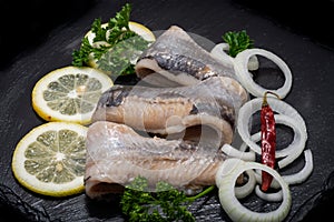 Salted Herring Fillets garnished with Yellow Onion, Lemon, Fresh Parsley and Dried Chili Pepper. Natural black stone background.