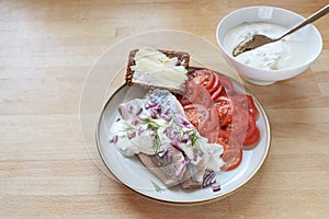 Salted herring fillets with sour cream sauce, red onions and dill on a plate with tomato slices and dark whole grainl bread,