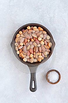 Salted roasted mixed nuts in black cast-iron pan on light stone background. Top view