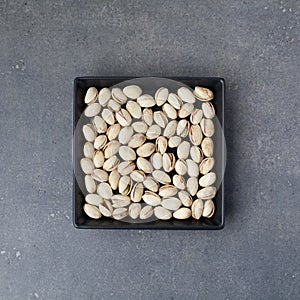 Salted roasted pistachios nuts with shell and salt in black small plate over gray rusty textured background. Top view