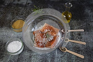 Salting salmon at home, fillet in a transparent bowl olive oil and spices in wooden spoons on the tabl