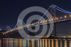 San Francisco Bay Bridge at night, lit up by yellow and blue lights, reflecting of the water in the Bay, long exposure