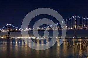 San Francisco Bay Bridge at night, lit up by yellow and blue lights, reflecting of the water in the Bay, long exposure