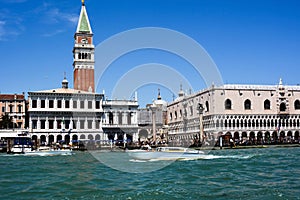 San Marco bell tower and surrounding buildings in Venice, Italy. Tourists are walking in front of them and a boat is sailing along