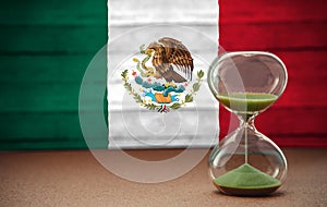 Sand running through the bulbs of an hourglass measuring the passing time in a countdown to a deadline, on mexico flag background