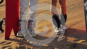 Sandboard, sandboarding. Feet closeup, two girls in boots with snowboard standing on the sand and preparing for the