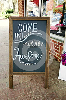 Sandwich board sign in front of a small business with Come In We`re Awesome message written in chalk