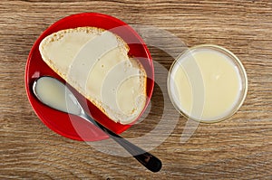 Sandwich with condensed milk, spoon in red saucer, transparent bowl with milk on wooden table. Top view