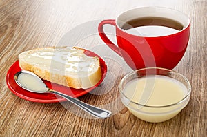 Sandwich with condensed milk, spoon in saucer, cup with tea, bowl with milk on wooden table