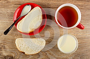 Sandwich with condensed milk, spoon in saucer, cup with tea, slice of bread, glass bowl with milk on table. Top view