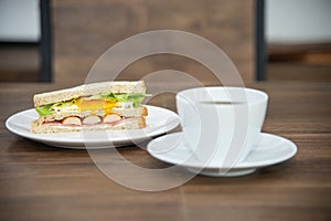 Sandwich and cup of coffee on white plate and wooden table, sandwich is made from bread, sausage, fried egg and vetgetable is