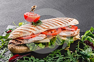 Sandwich with fillet grilled chicken, fresh vegetables, cheese and greens on black shale board over black stone background