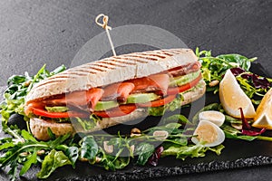 Sandwich with red fish, eggs, avocado, fresh vegetables and greens on black shale board over black stone background