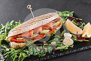 Sandwich with red fish, eggs, avocado, fresh vegetables and greens on black shale board over black stone background