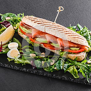 Sandwich with red fish, eggs, avocado, fresh vegetables and greens on black shale board over black stone background